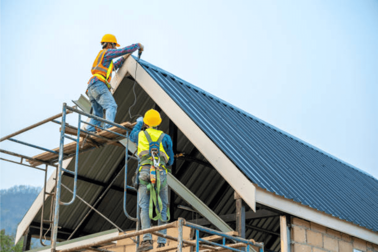 Two roofers in hard hats and safety harnesses fastening metal roof panels from scaffolding on a building under construction