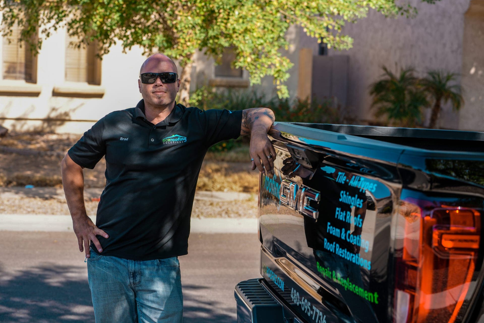 Eco Roofing Solutions production manager leaning on the tailgate of a branded truck lettered with the full service list — tile, shingles, flat roofs, foam and coating, roof restorations, repair and replacement