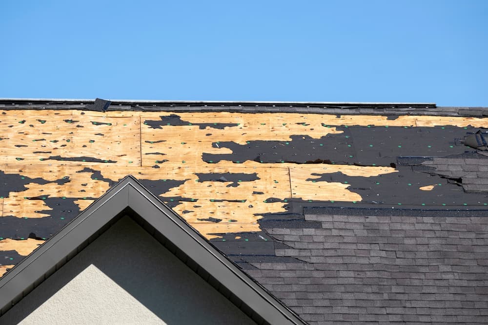 Storm-damaged roof with whole sections of shingles torn away, exposing the bare plywood deck underneath