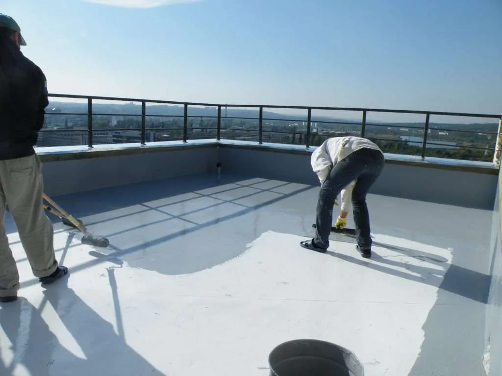 Two workers rolling a liquid waterproofing coat across a flat commercial roof deck behind a safety rail