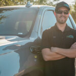 A smiling man named Mike, wearing an Eco Roofing polo shirt, cap, and sunglasses, stands with crossed arms in front of a light-colored GMC pickup truck.