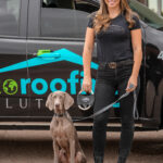 A woman in a black t-shirt and jeans standing next to a gray Weimaraner dog on a leash, with a black truck with a "Roofing Solutions" logo in the background.